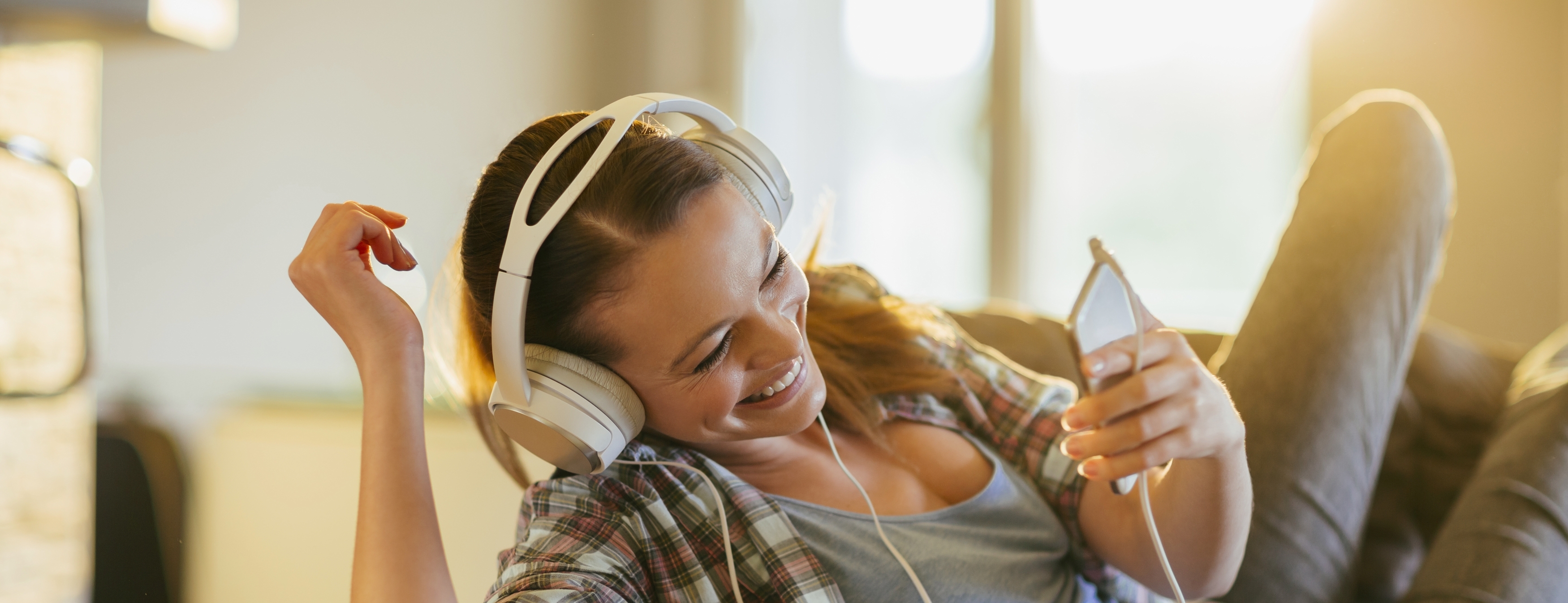 Picture of a young beautiful smiling woman relaxing at home in sofa. She is listening to music threw big headphones. In the background a window is letting in warm sunlight.